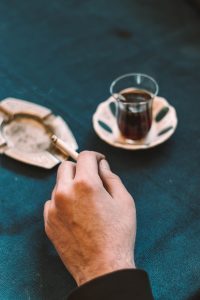 a person's hand ashes a cigarette into an ashtray next to the small brown drink they are drinking, which sits to the right in a small glass on a saucer