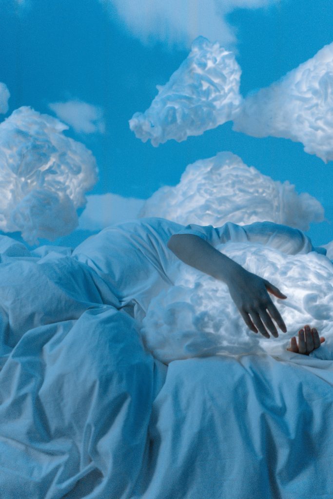 in a blue space with white fluffy clouds a sleeping person dressed in white grasps a fluffy flowing cloud like a pillow 